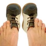 Imae of a pair of feet with tiny shoes on the big toes demonstrating incorrectly fitted shoes